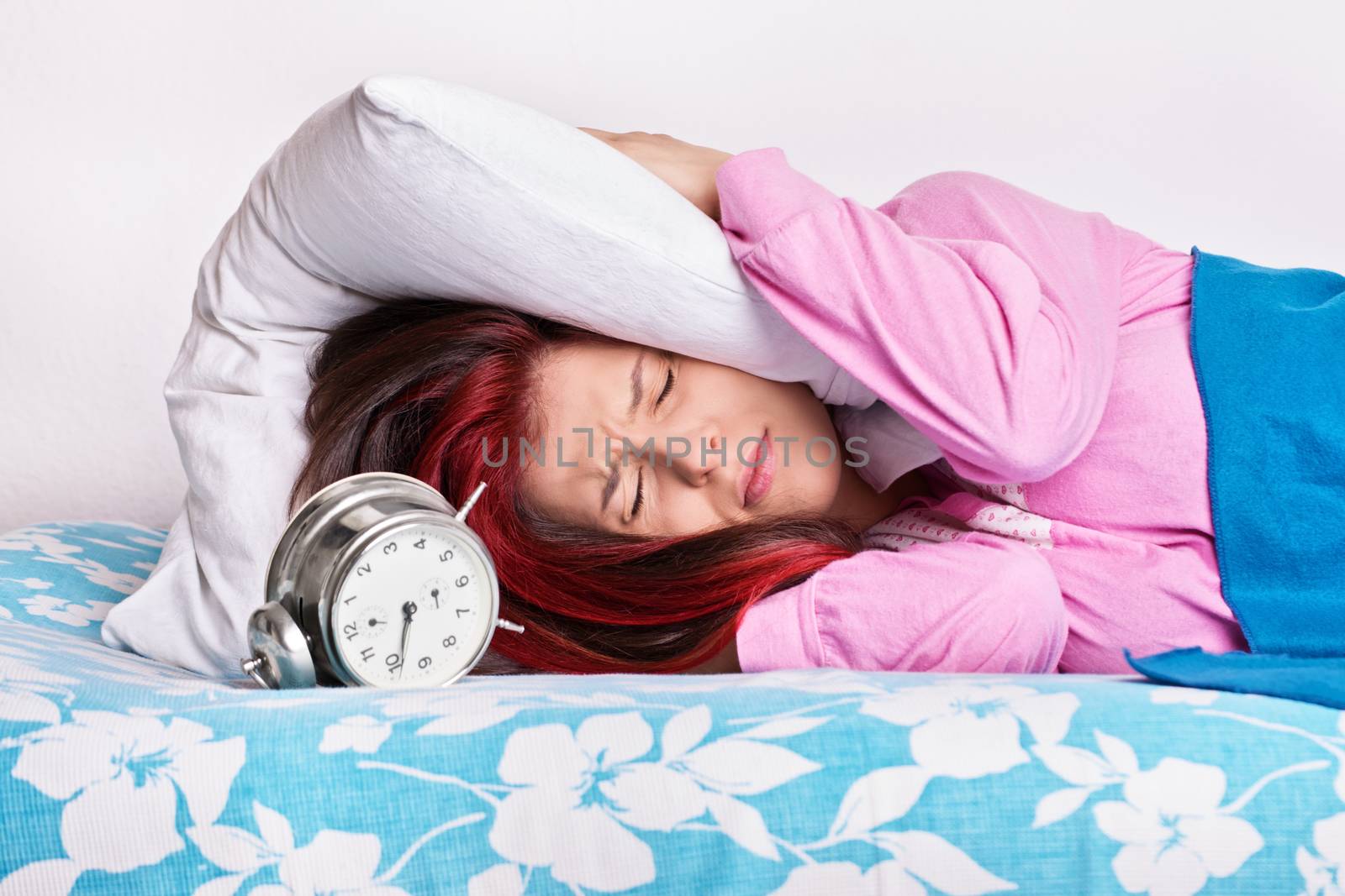Portrait of an annoyed young woman lying in bed covering her ears with a pillow due to the ringing of alarm clock in the morning. Student doesn’t want to wake up early for university or school. Oversleep, not enough sleep concept.