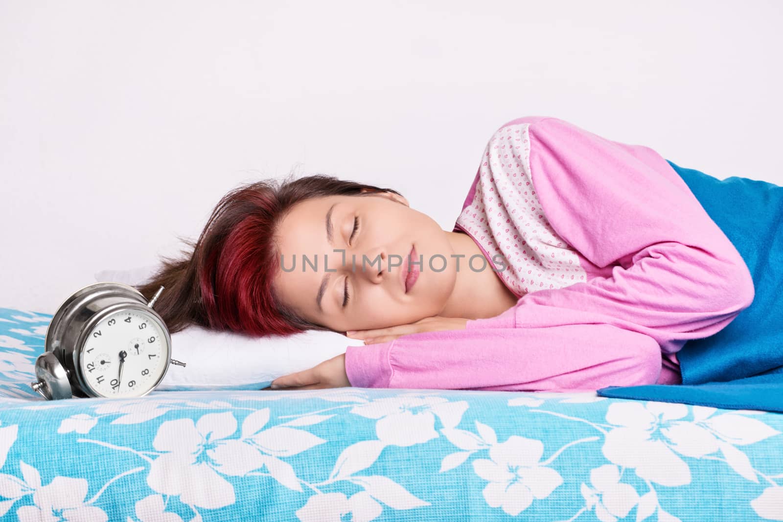 Beautiful young girl in pink pajamas calmly sleeping in her bed next to an old fashioned alarm clock. Healthy sleeping concept.