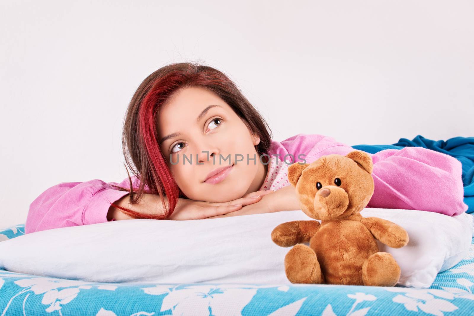 Beautiful young woman in pink pajamas lying in bed with her cute plush teddy bear, thinking of her dreams or a loved one.