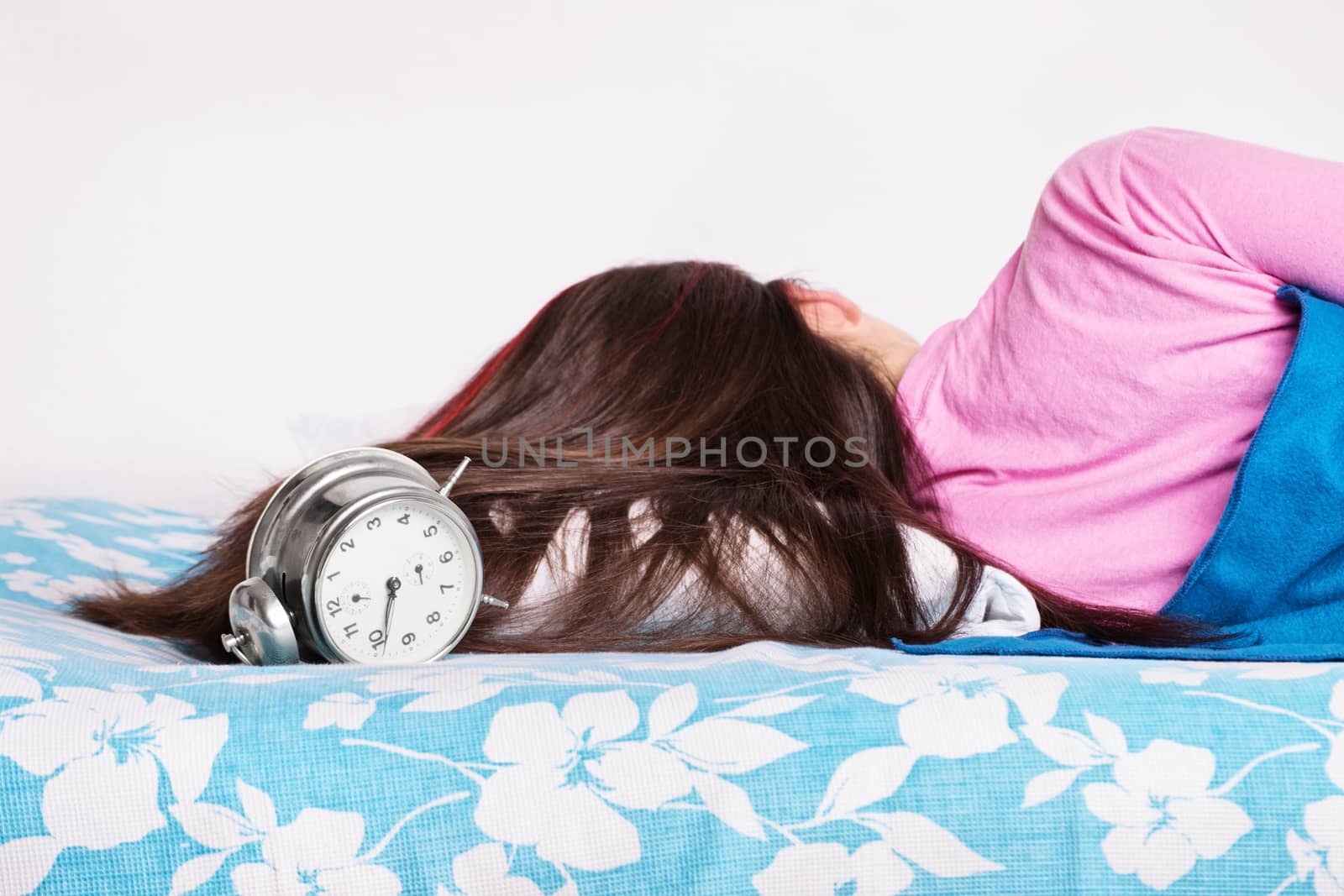 Beautiful young girl in pink pajamas calmly sleeping in her bed next to an old fashioned alarm clock, with her back turned to the camera. Healthy sleeping concept. Heavy sleeper concept.