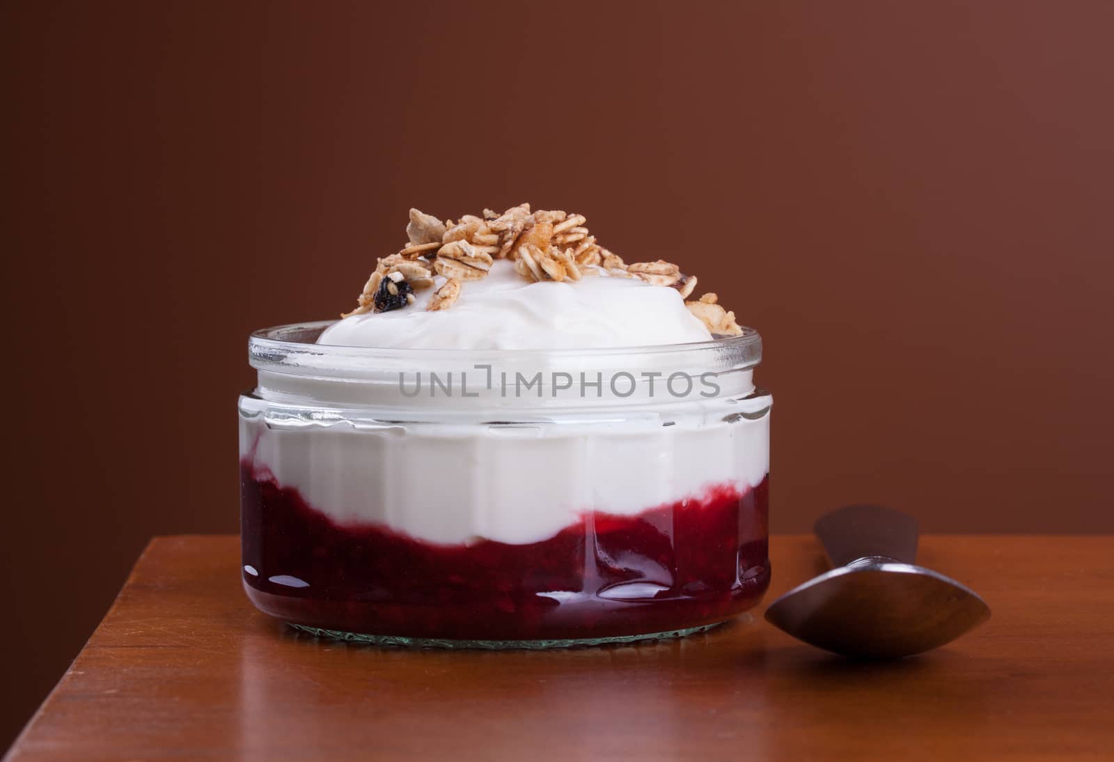 greek yogurt over raspberry jam and granola topping in a nice glass bowl place on a wood table near a silver and black spoon