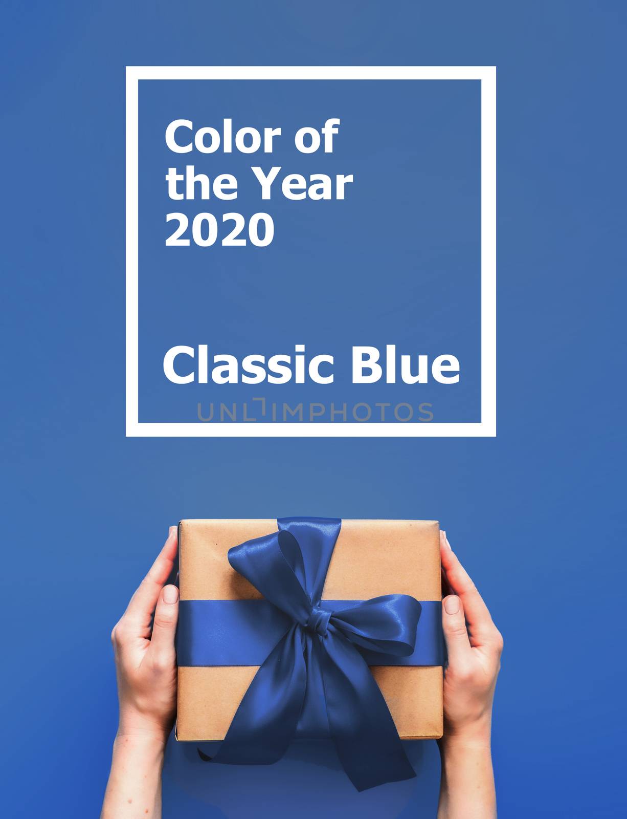 Female hands hold gift box on blue background with words Color of the Year 2020 Classic Blue. Gift box in craft wrapping paper with blue satin ribbon. Vertical.