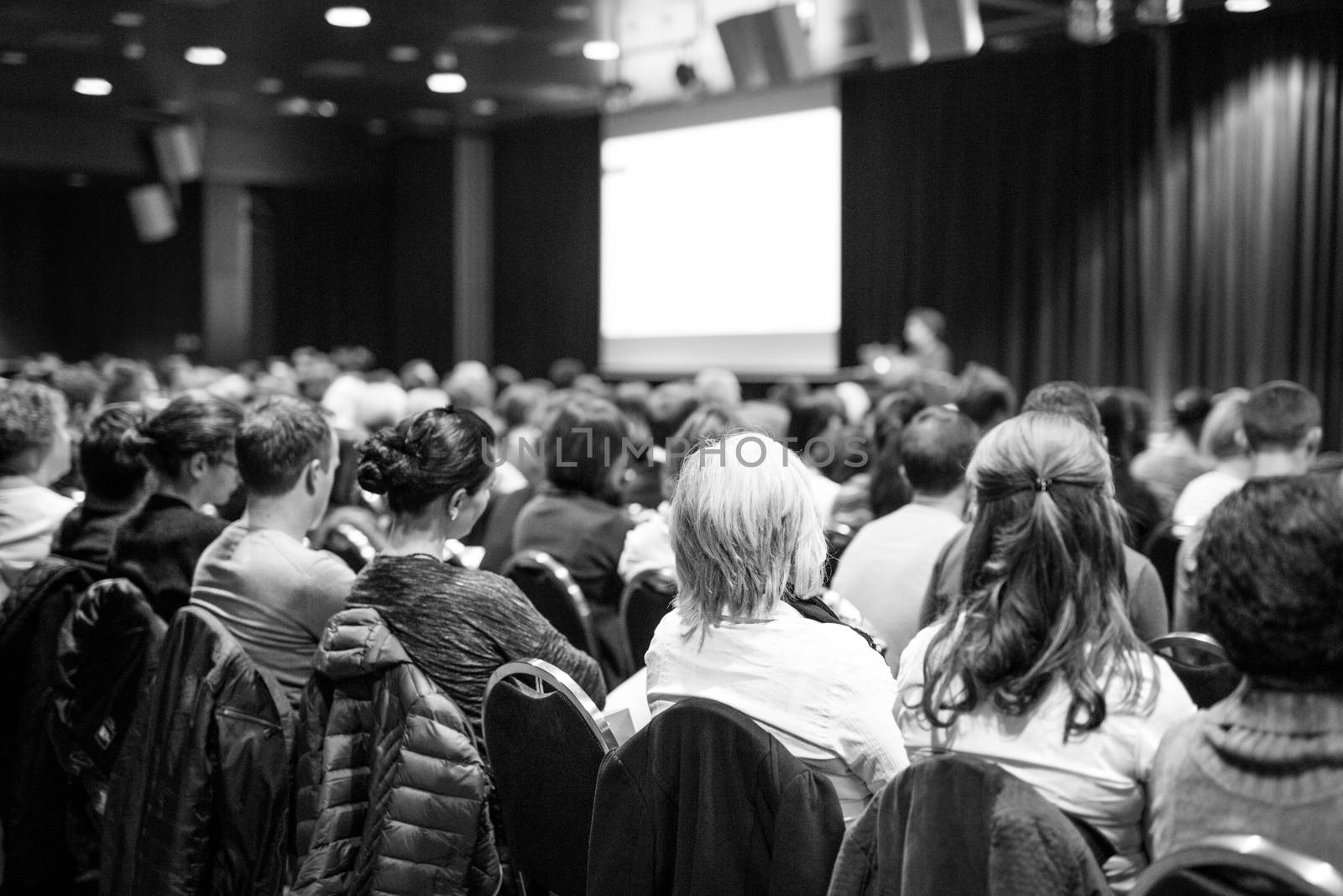 Business and entrepreneurship symposium. Speaker giving a talk at business meeting. Audience in conference hall. Rear view of unrecognized participant in audience. Black and white.