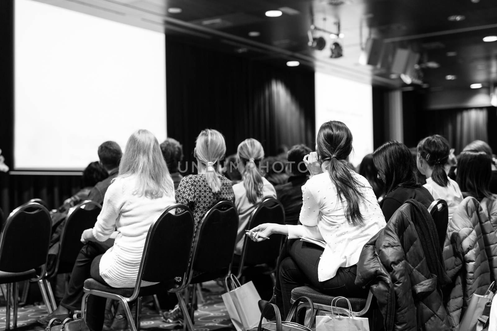 Business and entrepreneurship symposium. Speaker giving a talk at business meeting. Audience in conference hall. Rear view of unrecognized participant in audience. Black and white.