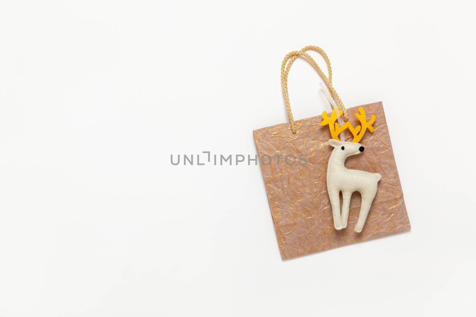 Golden Christmas gift bag, white felt toy deer with yellow horns on white background, copy space. Festive, New Year, sales, shopping concept. Horizontal, flat lay. Top view.