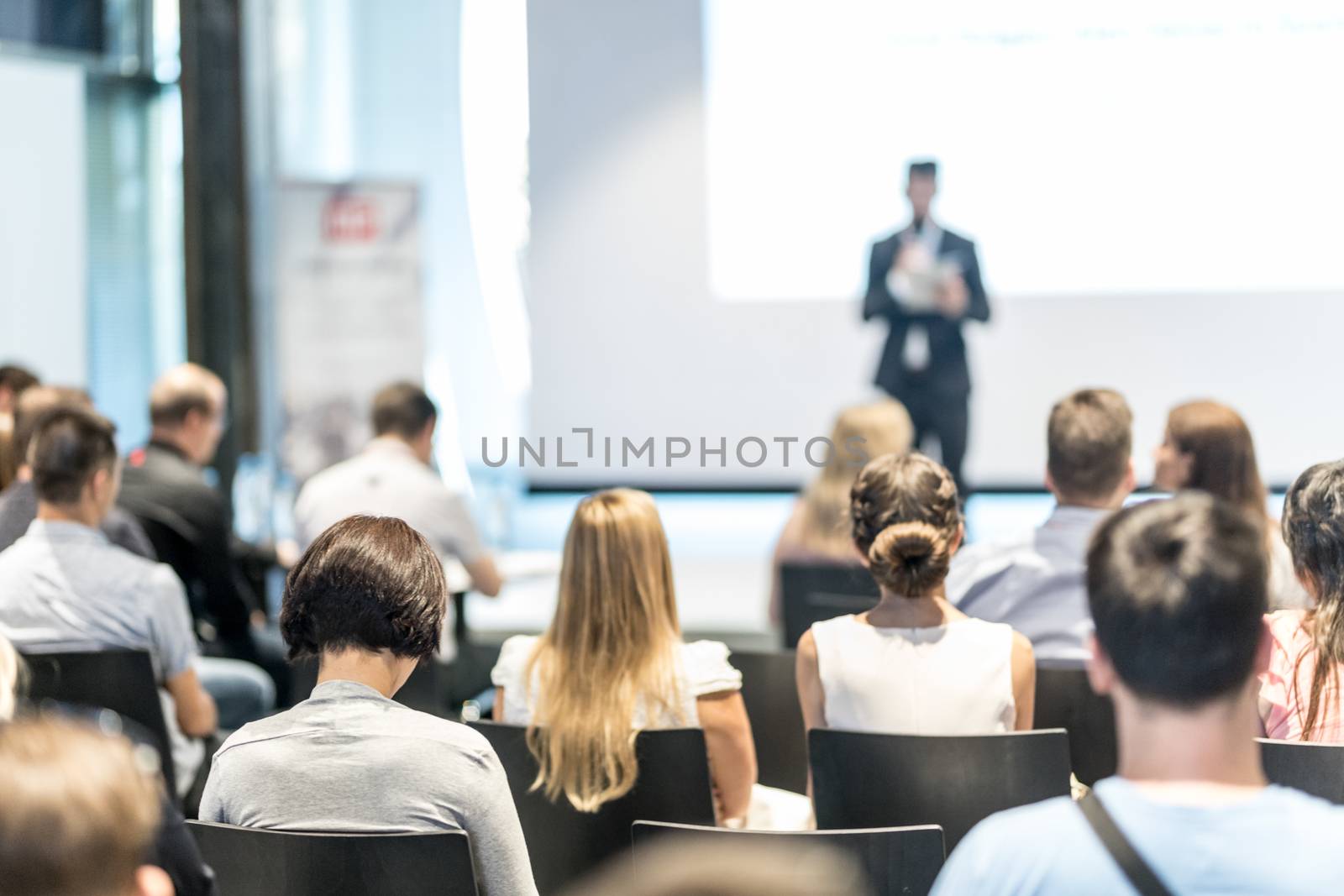 Business and entrepreneurship symposium. Speaker giving a talk at business meeting. Audience in conference hall. Rear view of unrecognized participant in audience.