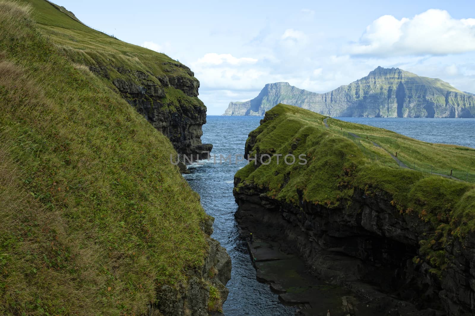 Direct view into the gorge / natural harbour of Gjogv with dramatic cloudy sky, lush green grass and turquoise water