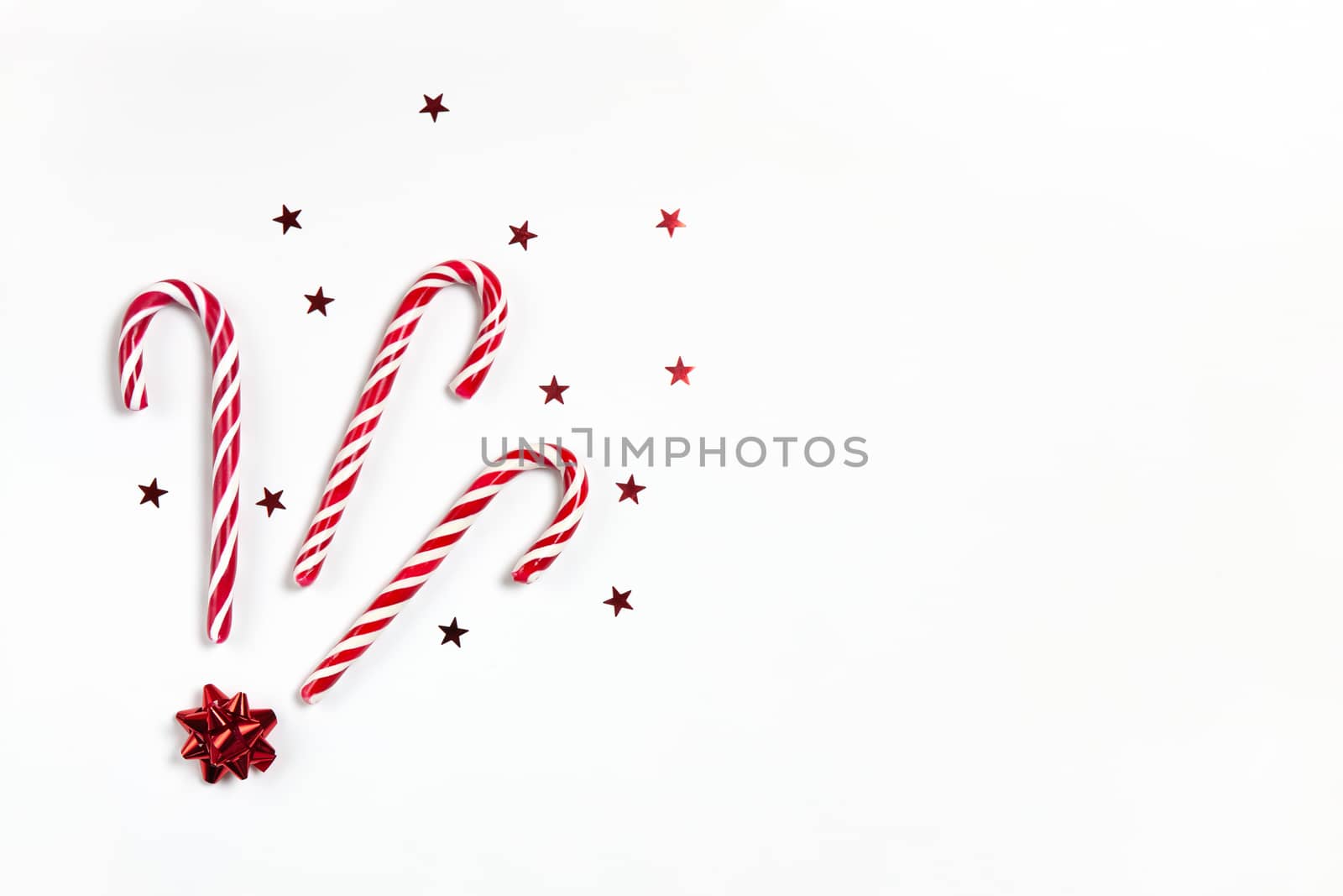 Christmas composition three caramel candy canes, confetti stars and red bow on white background with copy space. Festive minimal style flat lay. For greeting card, invitation. Horizontal.