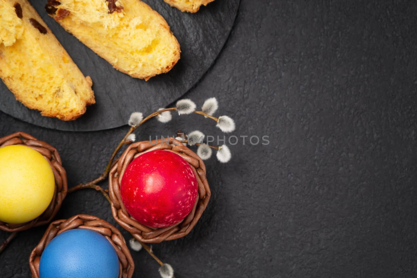 Painted Easter eggs in wicker coasters, pieces of Easter cake on a cutting board on a dark table - traditional Easter breakfast, top view, flat lay.