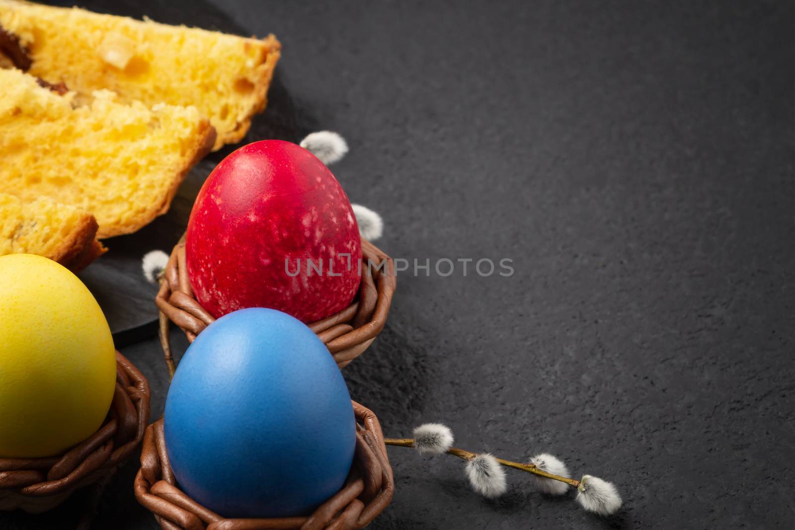 Painted Easter eggs in wicker coasters, pieces of Easter cake on a cutting board on a dark table - traditional Easter breakfast.