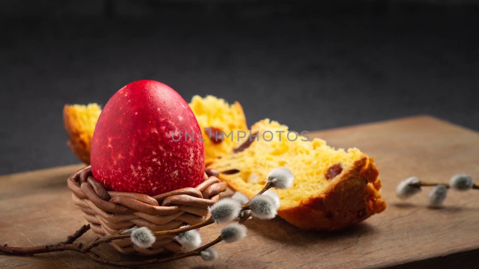 Red Easter egg in wisker stand and slice of Easter cake on a cutting board on a dark table - traditional Easter breakfast.