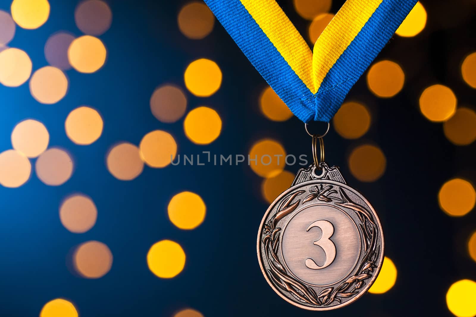 Third placed runner up bronze medallion on a ribbon hanging against a blue background with golden bokeh
