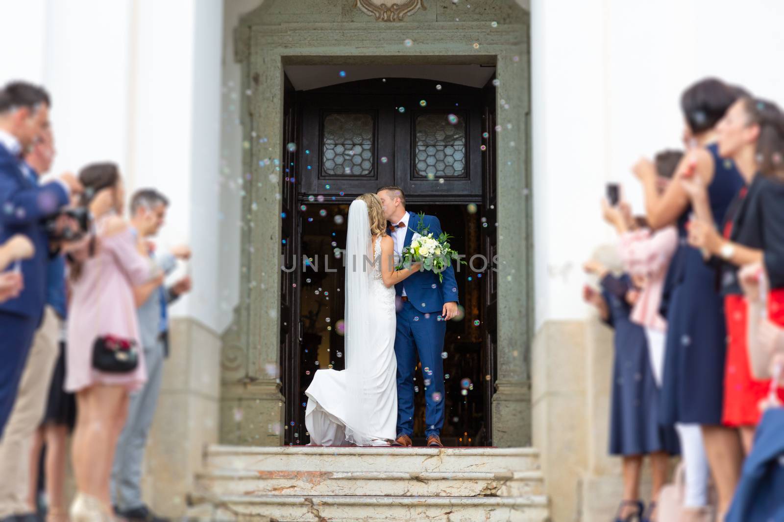 Newlyweds kissing while exiting the church after wedding ceremony, family and friends celebrating their love with the shower of soap bubbles, custom undermining traditional rice bath.