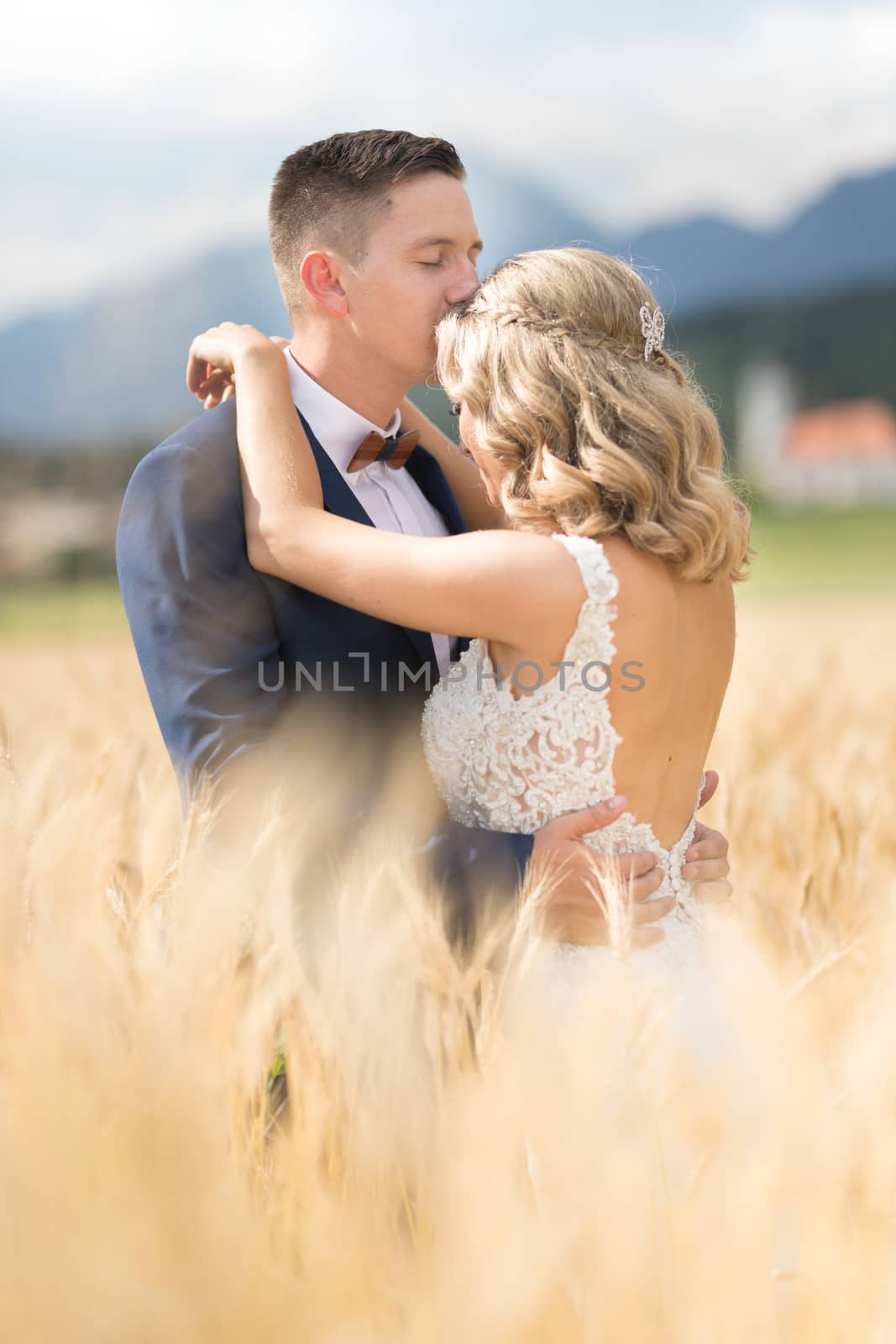 Groom hugging bride tenderly and kisses her on forehead in wheat field somewhere in Slovenian countryside. Caucasian happy romantic young couple celebrating their marriage.
