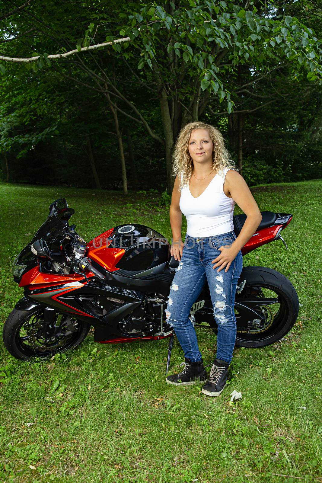 Twenty something blond woman, wearing a white tank to and jeans, standing in front of her motocycle