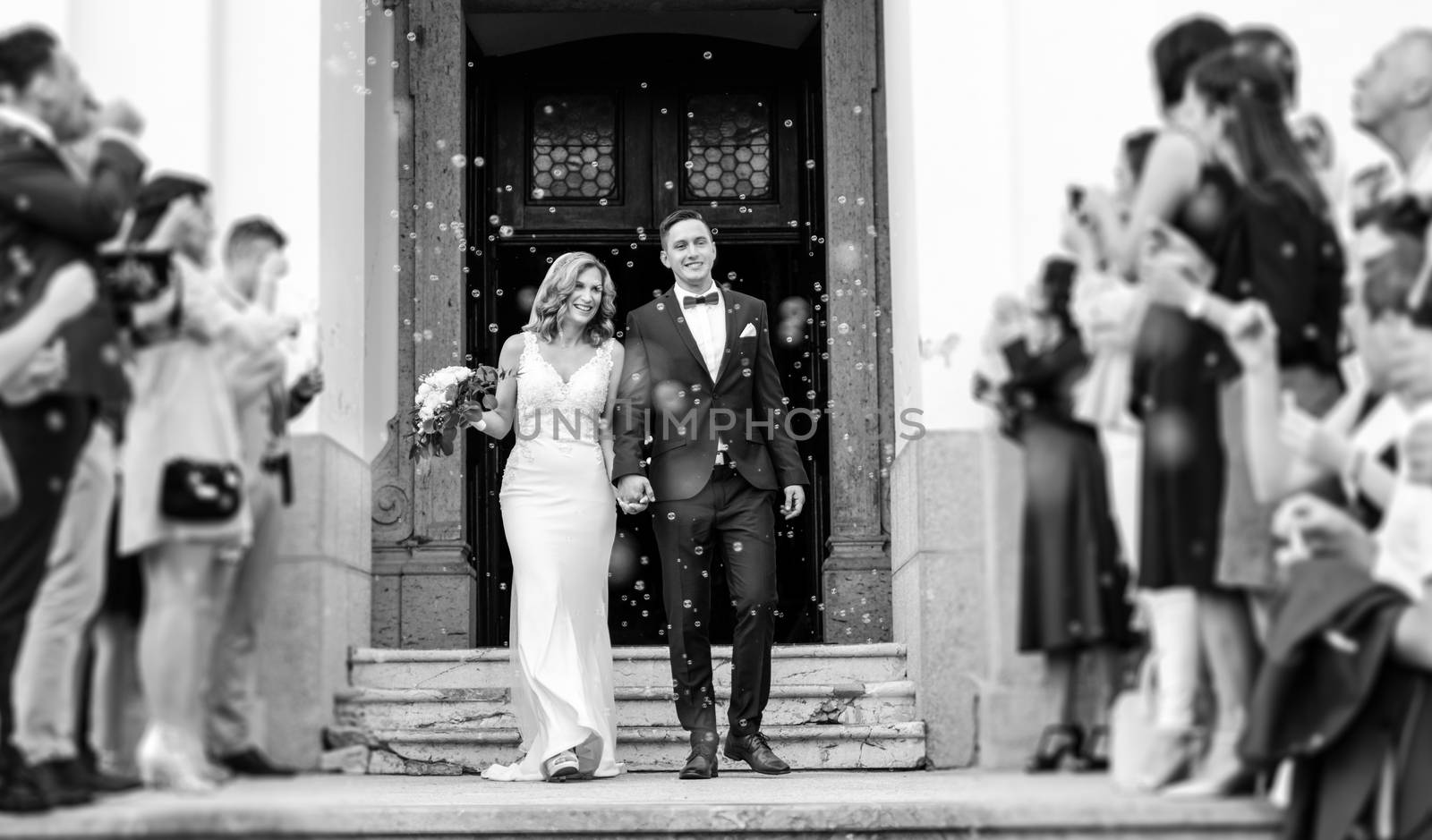 Newlyweds exiting the church after the wedding ceremony, family and friends celebrating their love with the shower of soap bubbles, custom undermining traditional rice bath by kasto