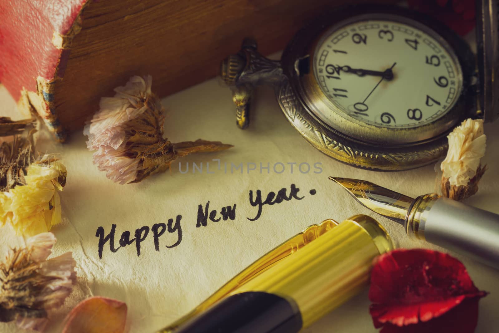 Vintage brass pen writing happy new year greetings on old paper and old book stacked on wooden table. Decoration by pocket watch and dry flower around it.