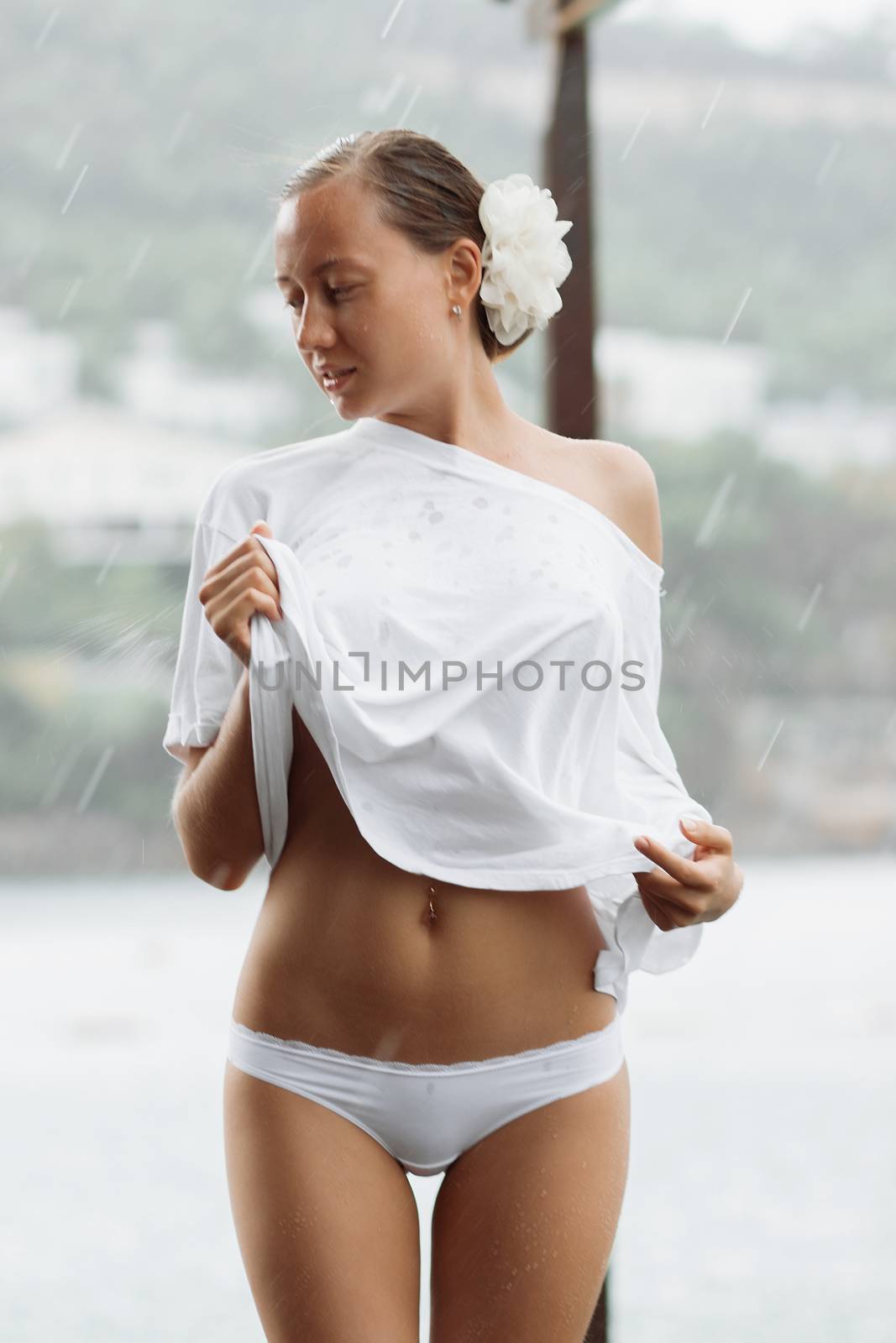 Attractive young female in while blouse and panties embracing herself and keeping eyes closed while standing near water on windy day