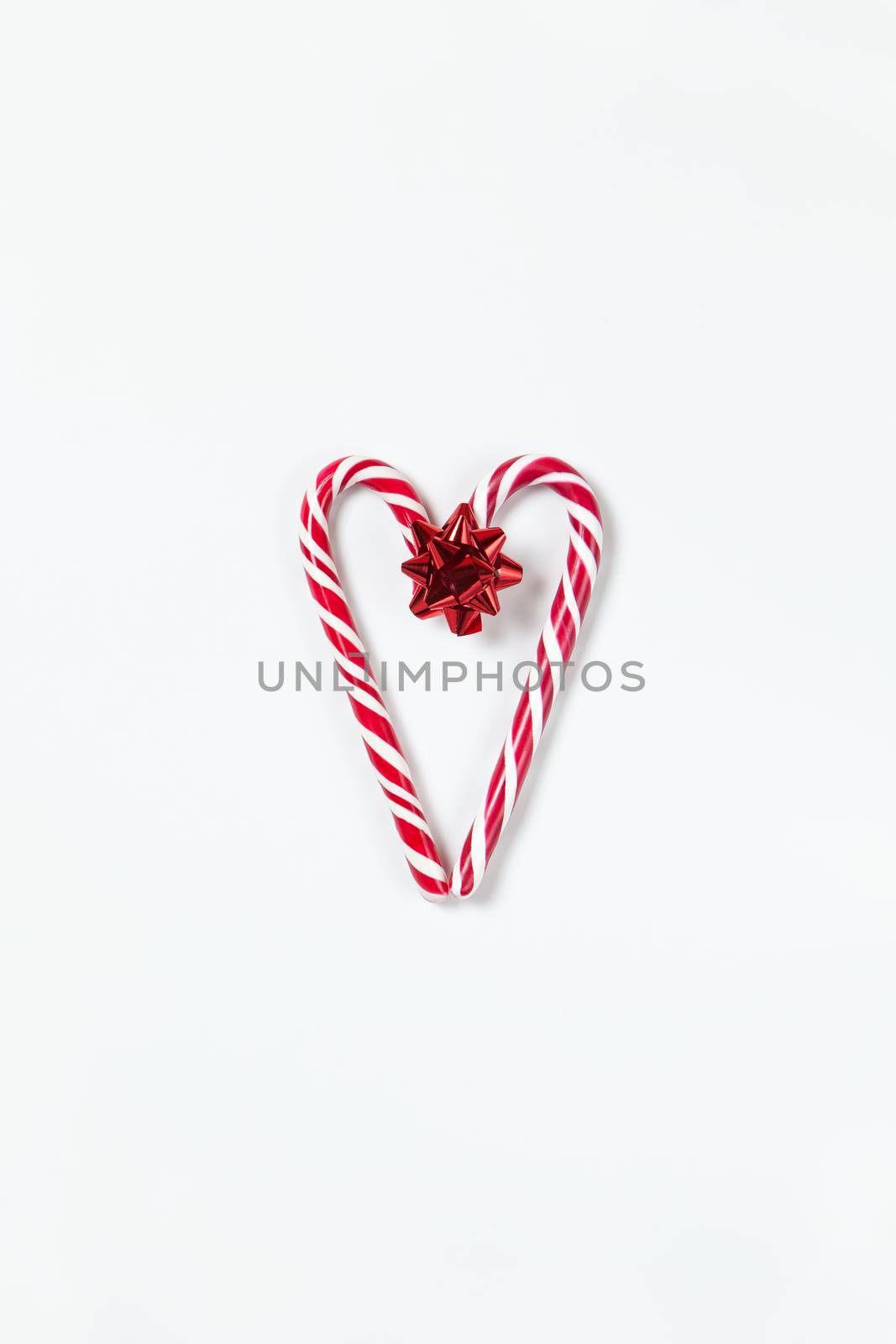 Christmas composition, heart made of two caramel candy canes and red bow on white background. Festive minimal style flat lay. For greeting card, invitation, social media. Vertical orientation.