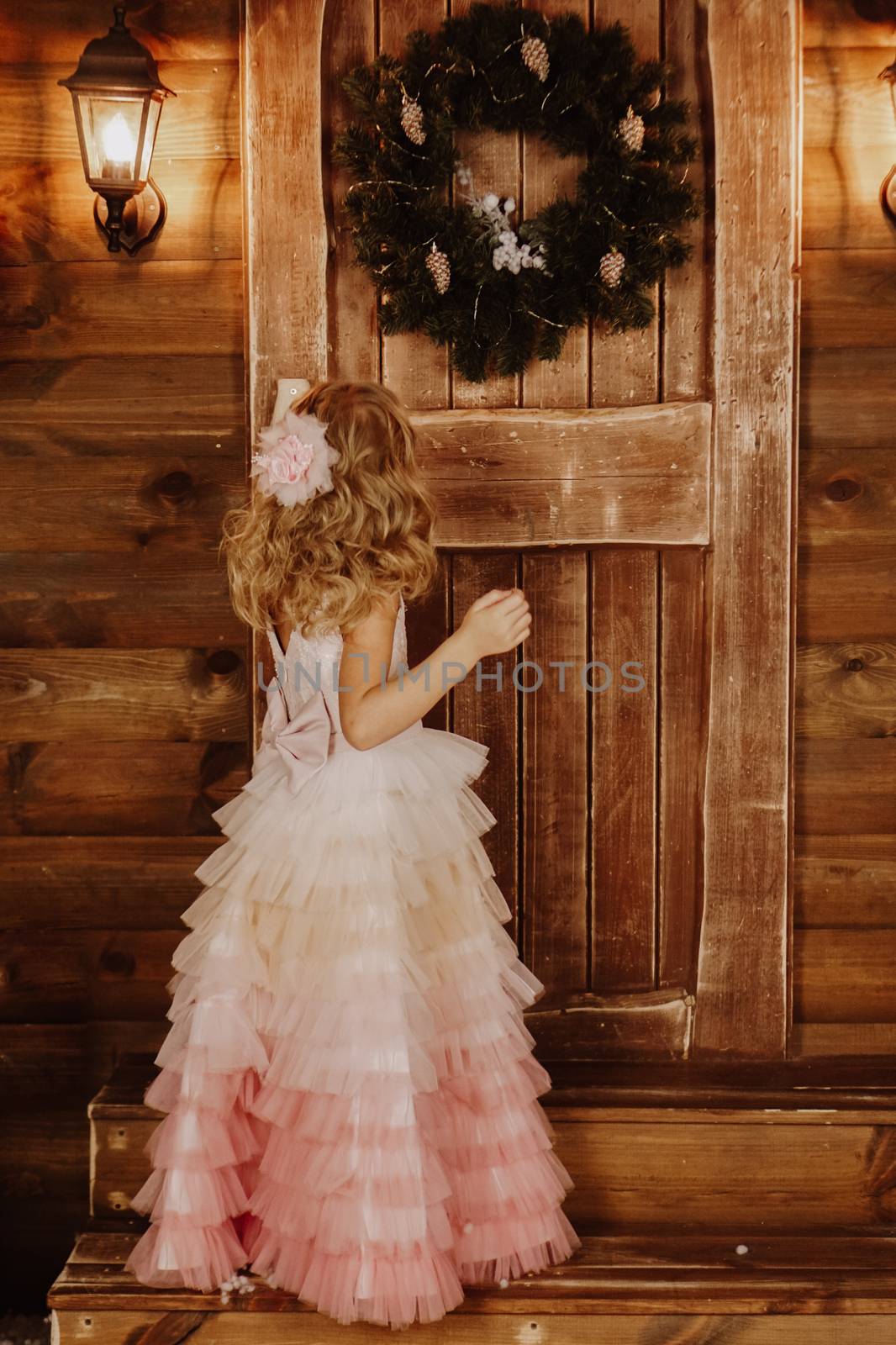 Little girl in pink dress stands in front of wooden door with Christmas wreath by natali_brill