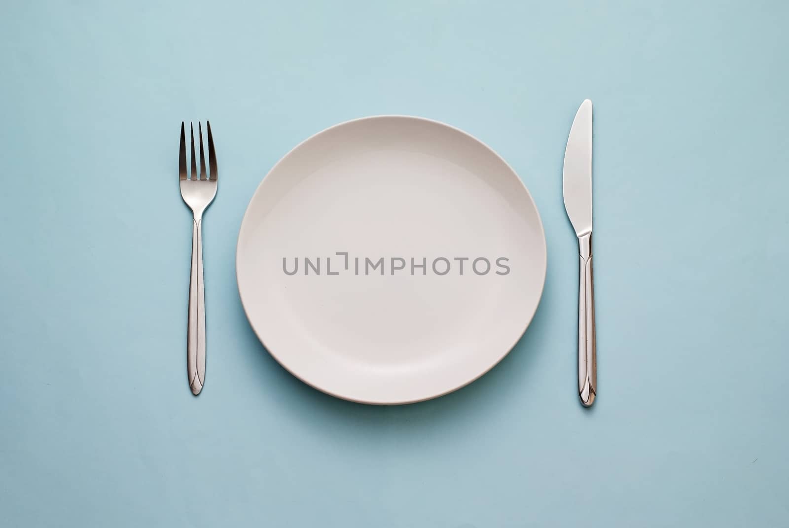Clean empty white plate with knife and fork on a blue background conceptual of eating, food and catering