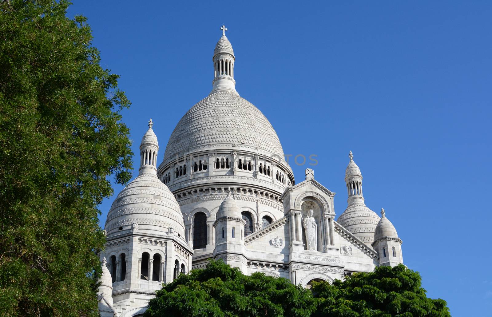 Dome of the Sacre Coeur basilica in Paris rises above trees against a blue sky, showing detailed architecture