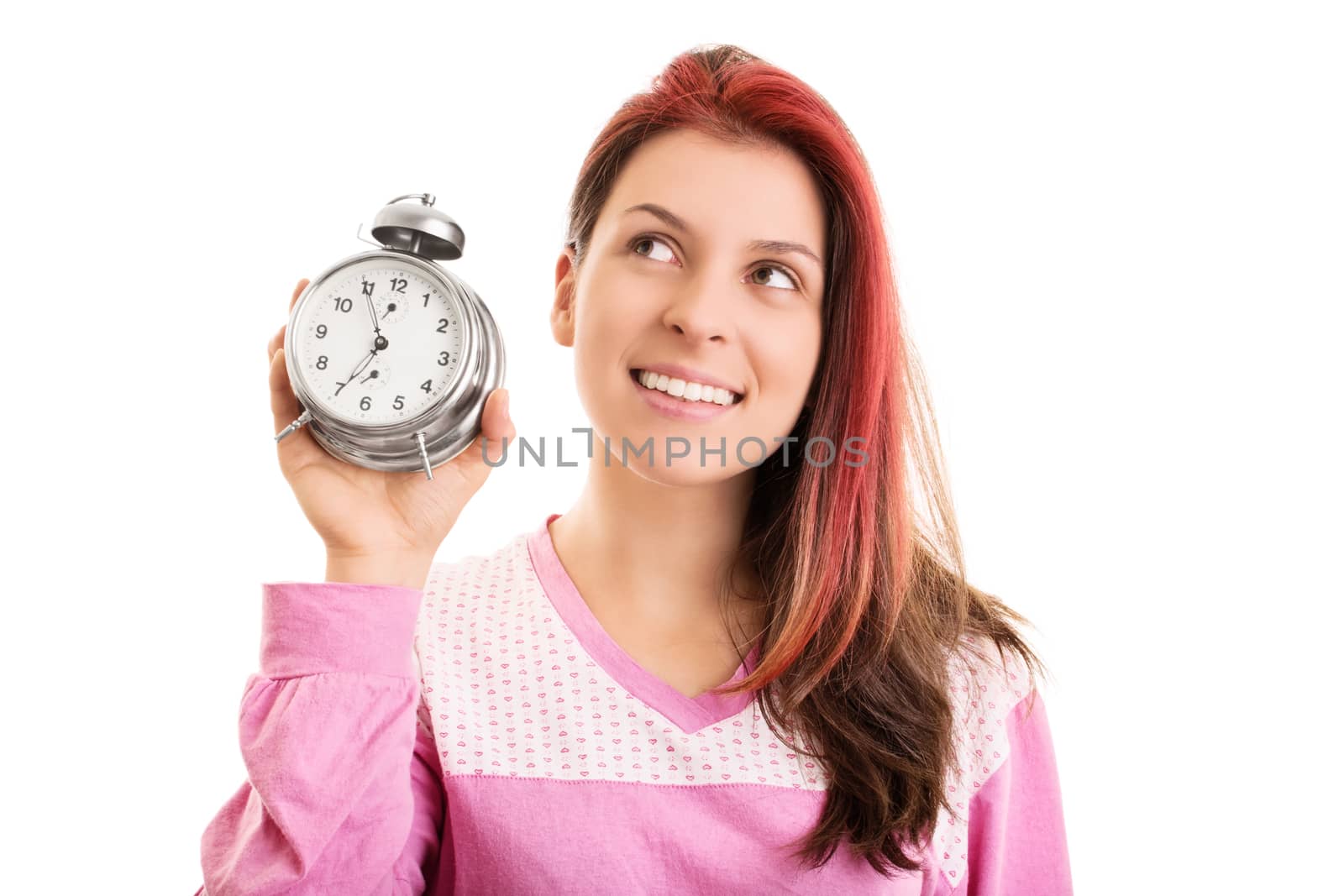 Early morning concept. Close up of a beautiful smiling young woman in pink pajamas holding an alarm clock looking up, isolated on white background.