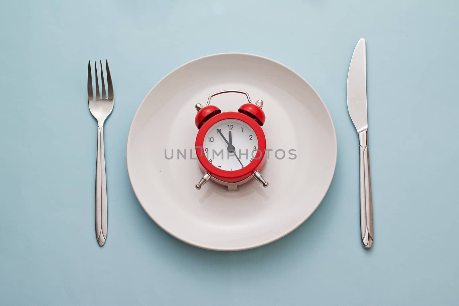Red alarm clock on a clean white dinner plate with knife and fork alongside on a blue background viewed from above