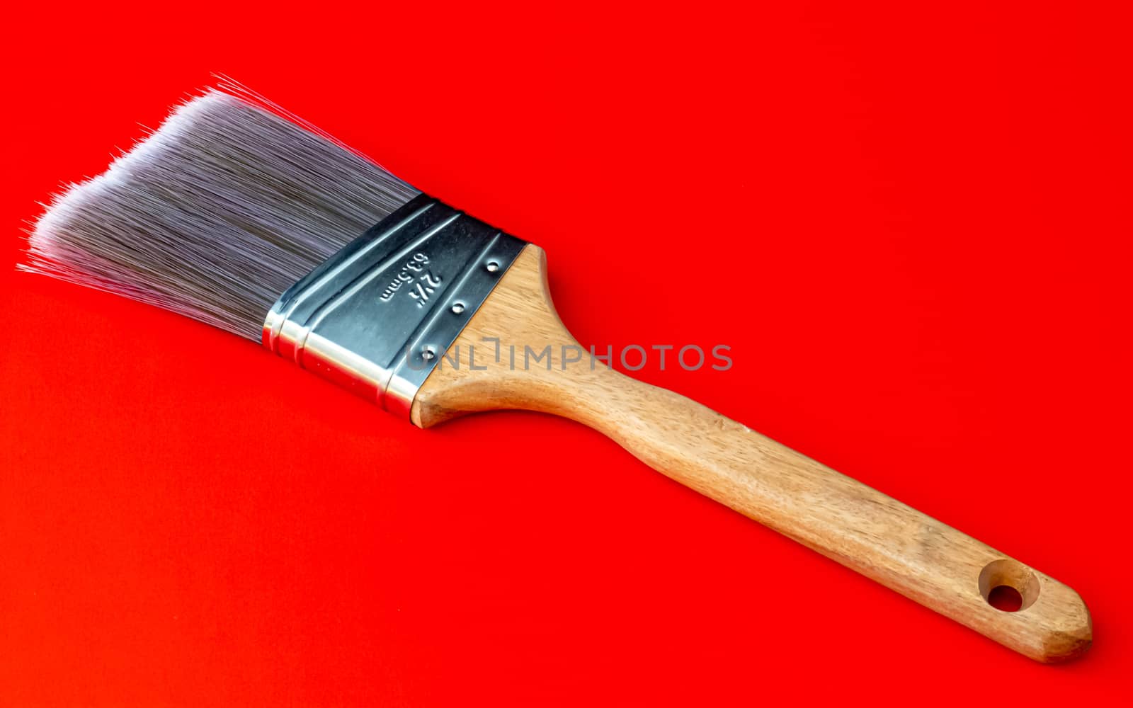 A large format paint brush sits on a solid red background. The paint brush has a wooden handle and polyester/nylon bristles emerging from a shiny stainless steel ferrule.
