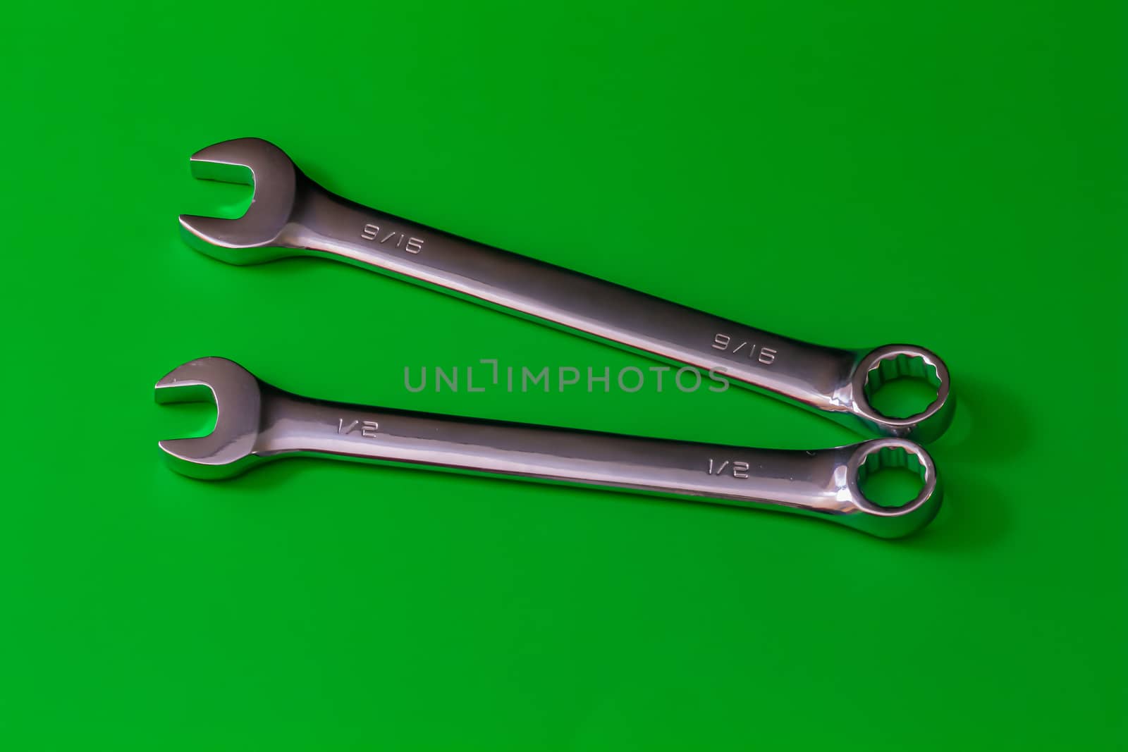 A pair of stainless steel wrenches, combination spanners with both an open end and a box end, lie on a solid green background. their sizes are engraved as fractions of an inch at 1/2" and 9/15".