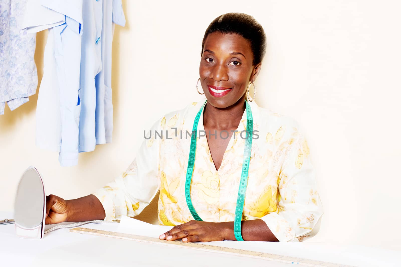 Young smiling seamstress holding an iron in front of her work table.