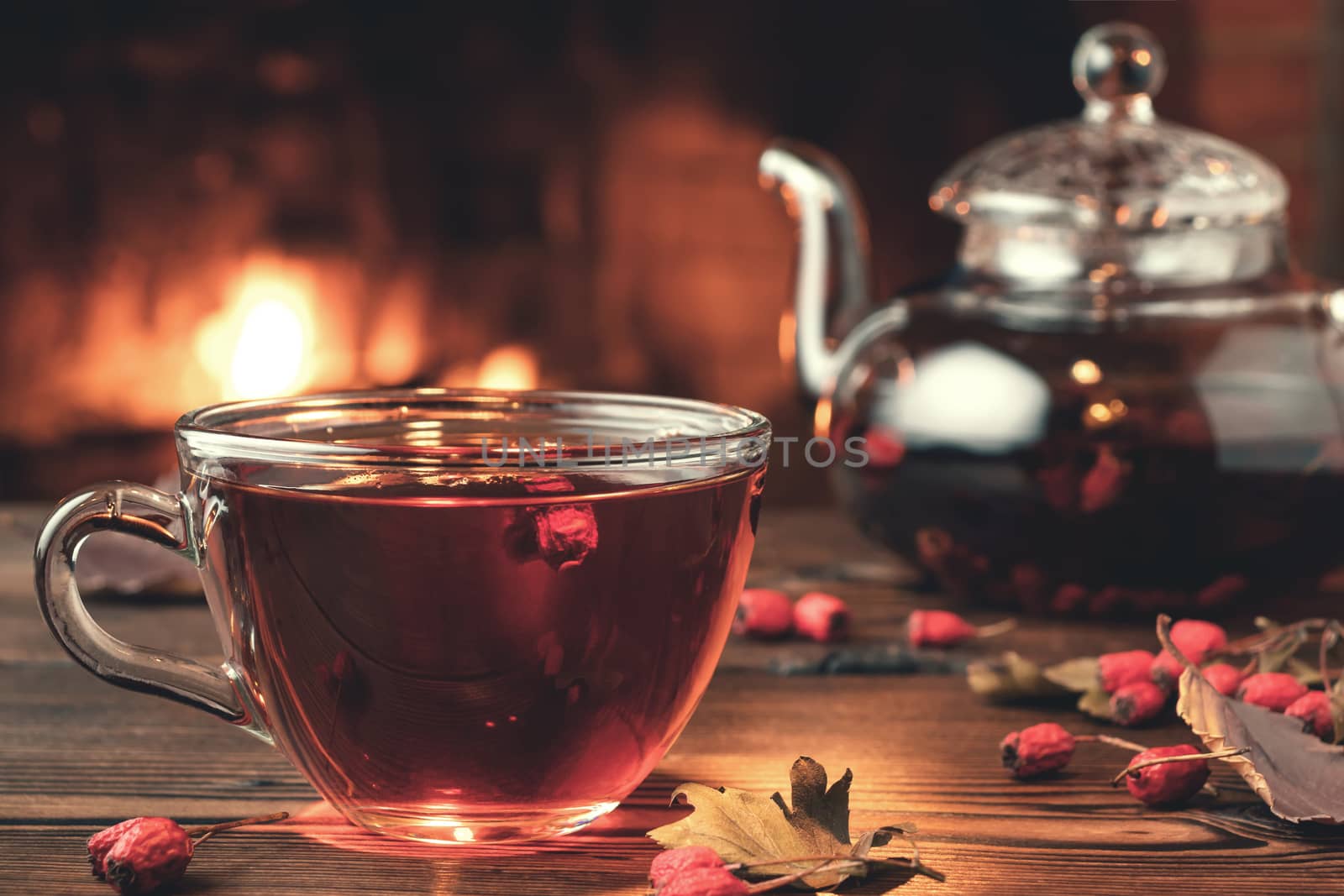 Tea with hawthorn in a glass cup and teapot on a wooden table in a room with a burning fireplace, closeup.