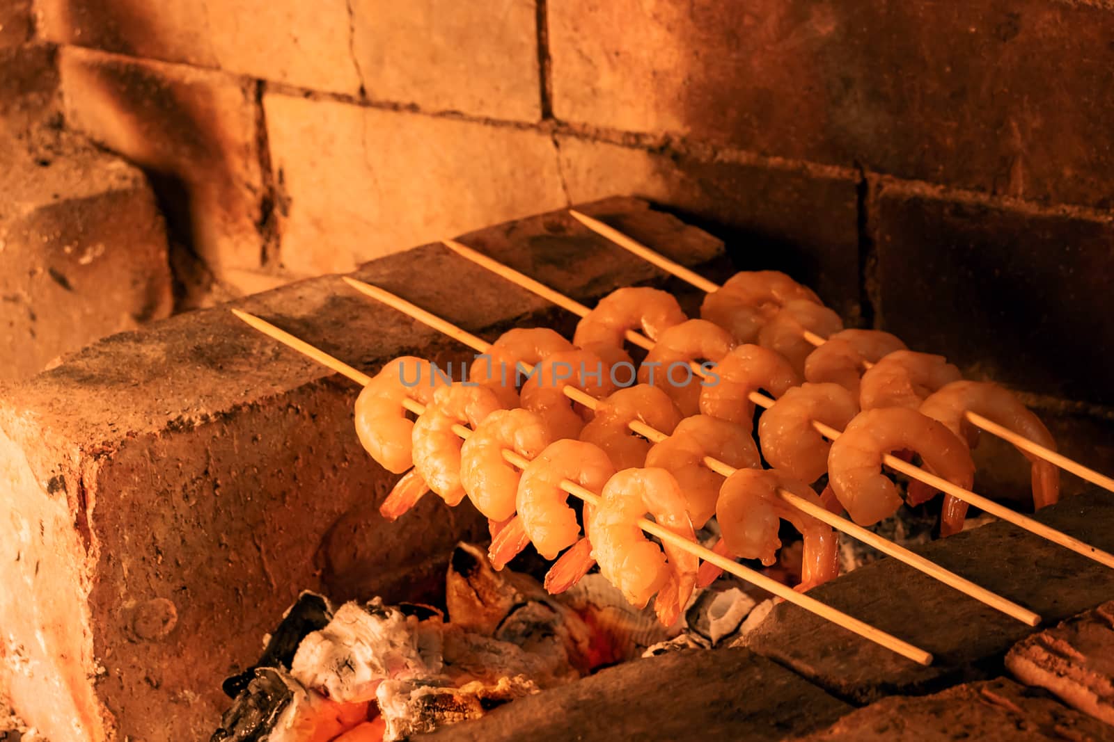 Shrimp frying on bamboo skewers on hot coals in a fireplace.