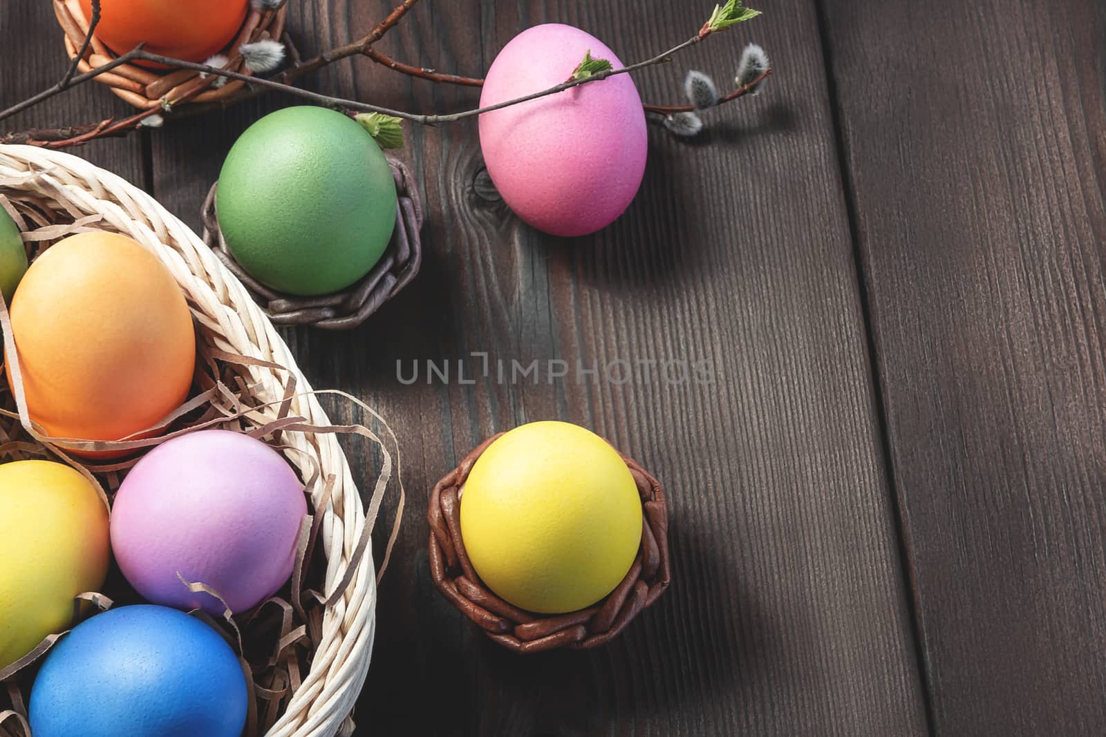 Easter composition - several colored eggs in a basket and on a dark wooden table with willow twigs, place for text, copy space.