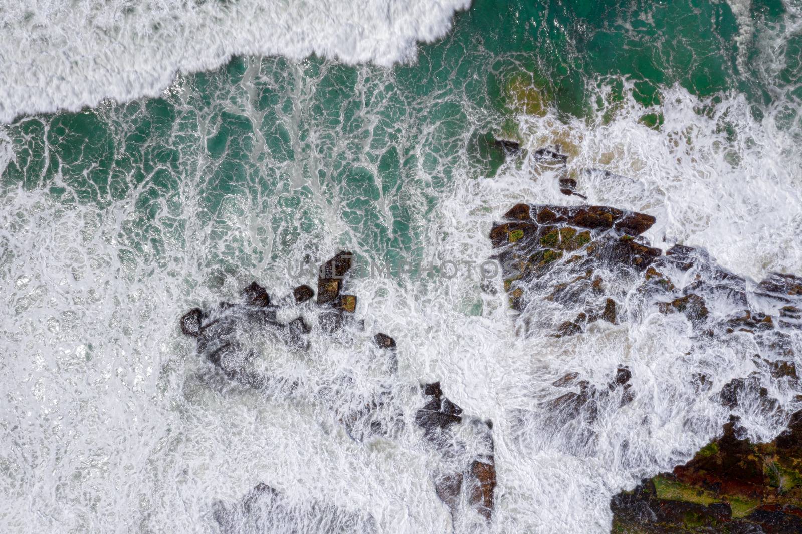 Wave action over rocks, motion in water. Rocks eroded into patterns, some with green moss and algae. Aerial perspective