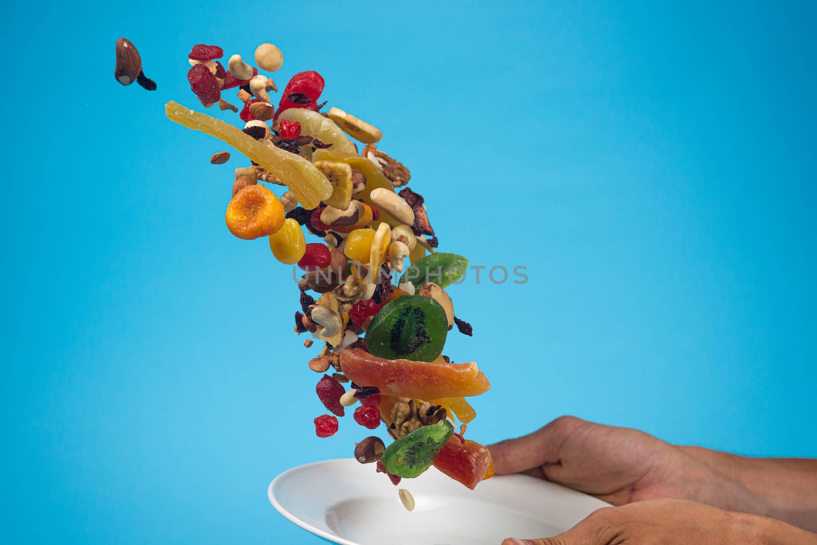 Male hands holding an empty white bowl on blue background. Candied fruits and nuts flying above the bowl. Stock photo of nutrient and healty food. Conceptual photo of vegan and vegetarial food and meal.