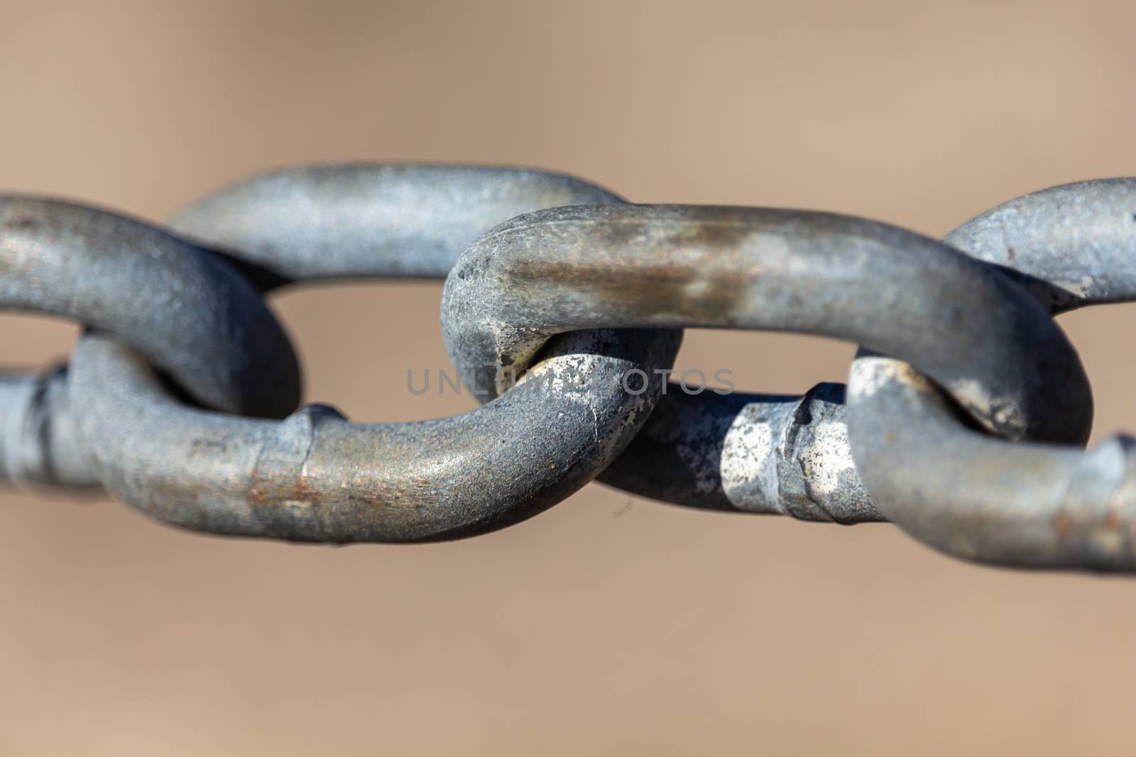 The links of a steel chain are seen up close. The chain is held up taut and viewed against a blurry beige background. Some wear and weathered detail is seen, showing its outdoor exposure.