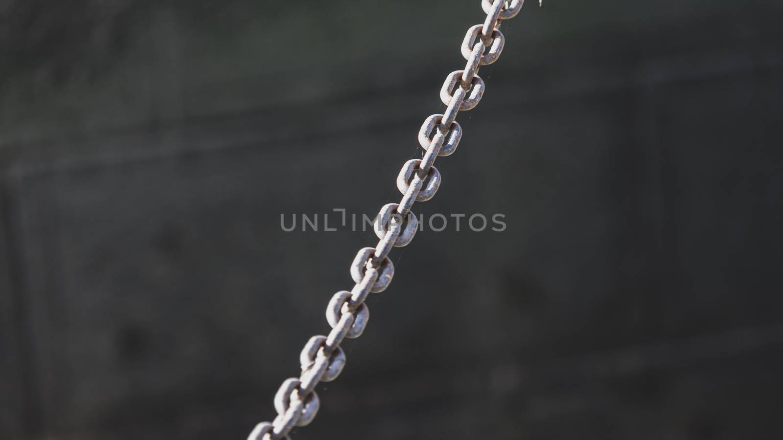 A metal chain made of steel is suspended across an open area, and viewed such that it runs diagonally across the center of the frame. Darkened gray stone bricks are seen out of focus behind it.