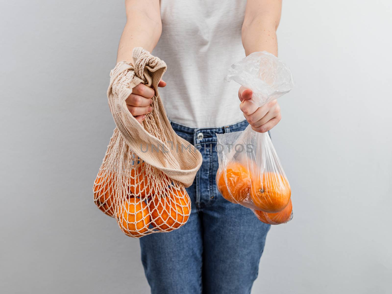 Oranges fruits in plastic bag and oranges in textile mesh bag in female hands near white wall background. Zero waste, food waste concept.