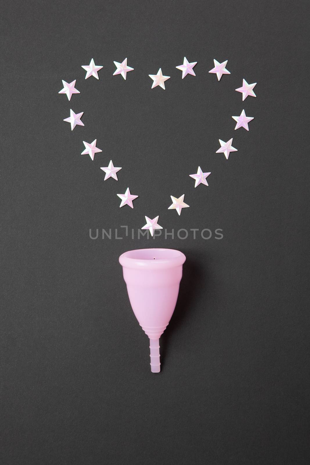 Pink menstrual cup on black background with heart, copy spase. Concept possibility of comfort use of cup at night, zero waste, savings, minimalism. Feminine hygiene product, flat lay. Vertical.