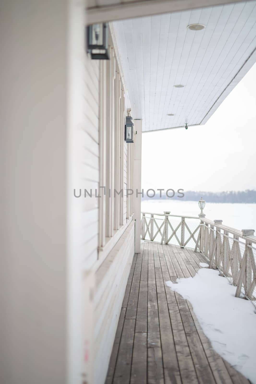 white snow on the wooden vintage open deck, old peeling paint light milk color, steel and copper lanterns hanging on the wall, snowy winter, snow lies on the river and the far shore of the earth is vi