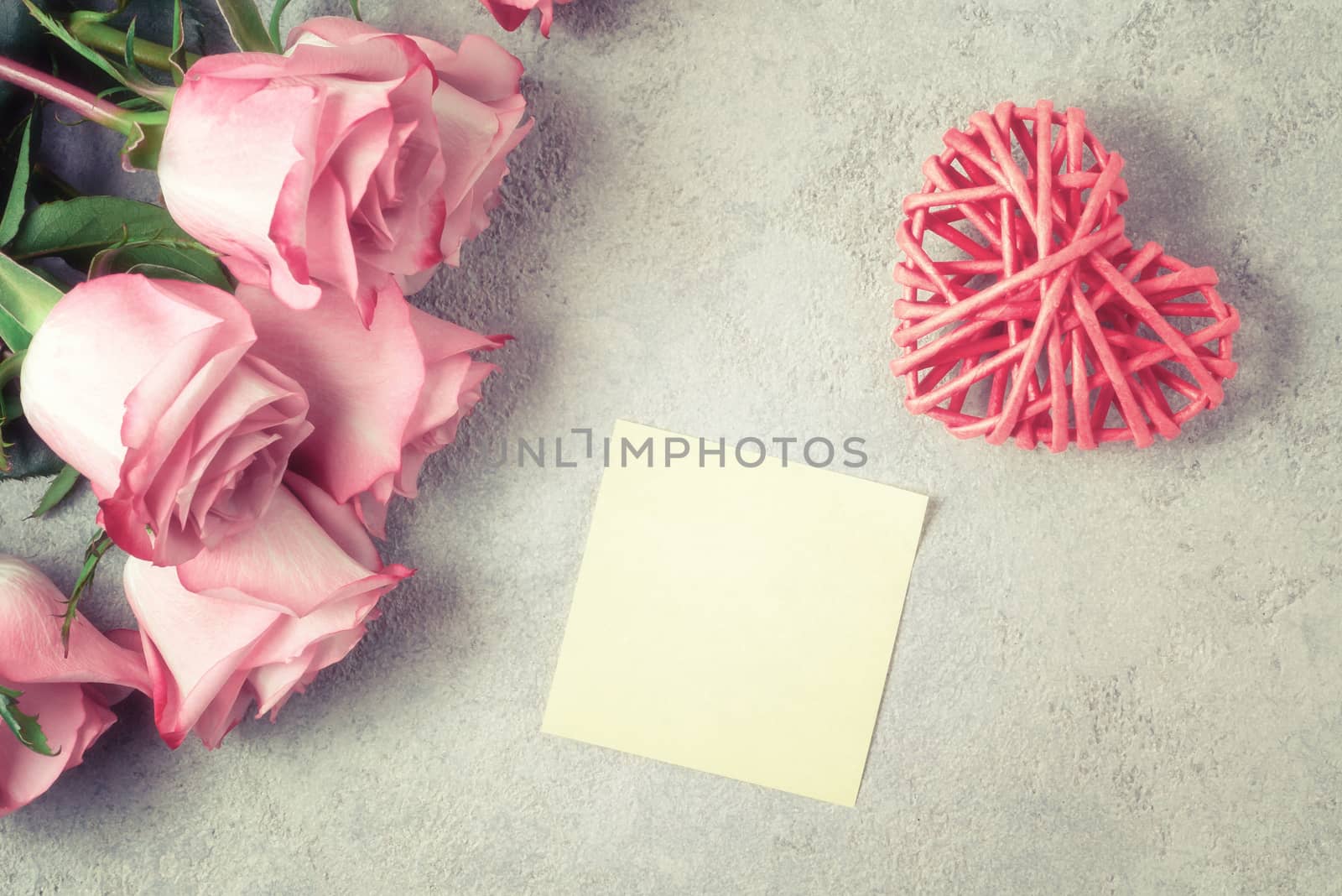 Flower arrangement - a bouquet of pink roses, a wicker heart and an empty sheet for a note on a concrete surface.