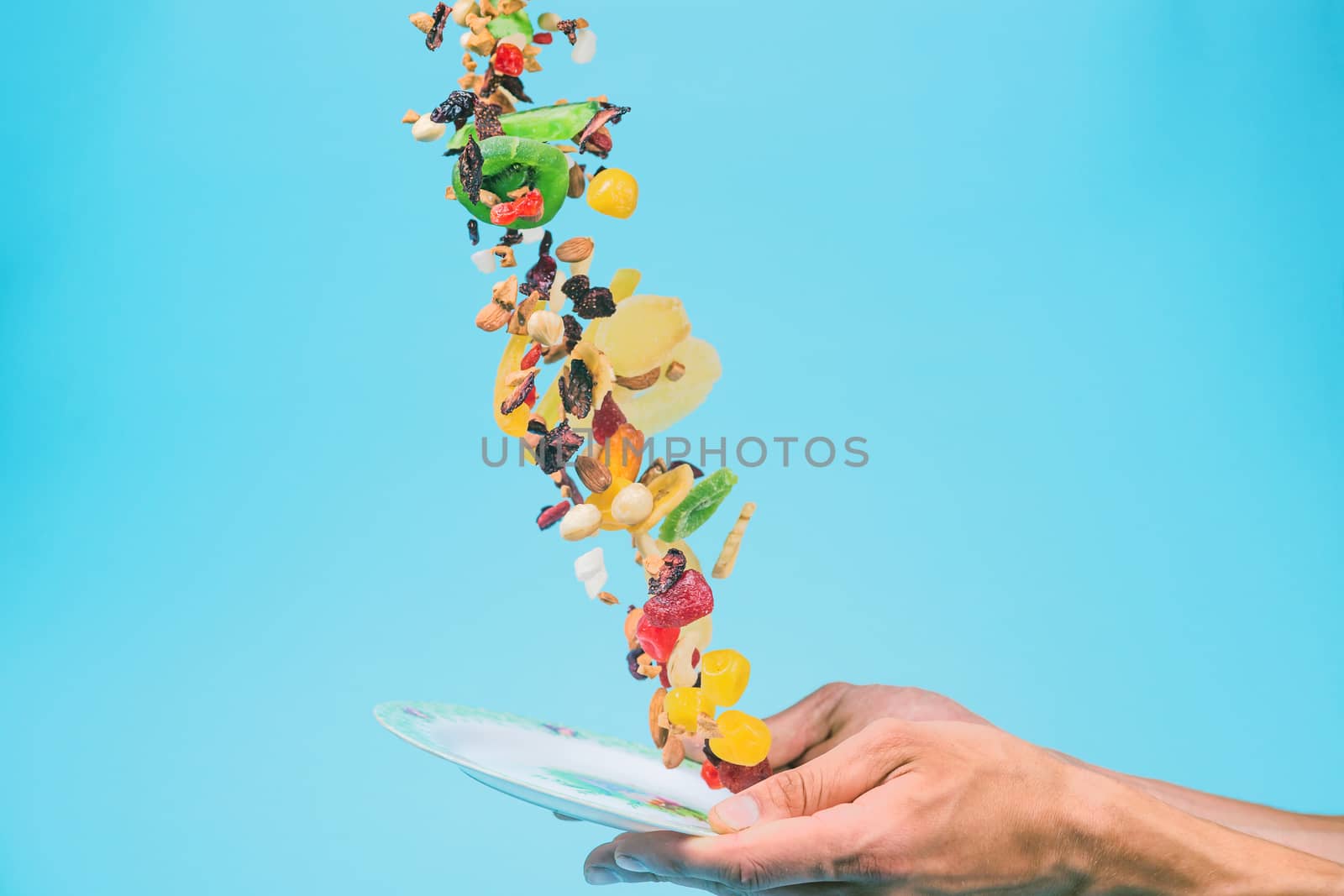 Male hands holding an empty white bowl on blue background. Candied fruits and nuts flying above the bowl. Stock photo of nutrient and healty food. Conceptual photo of vegan and vegetarial food and meal.