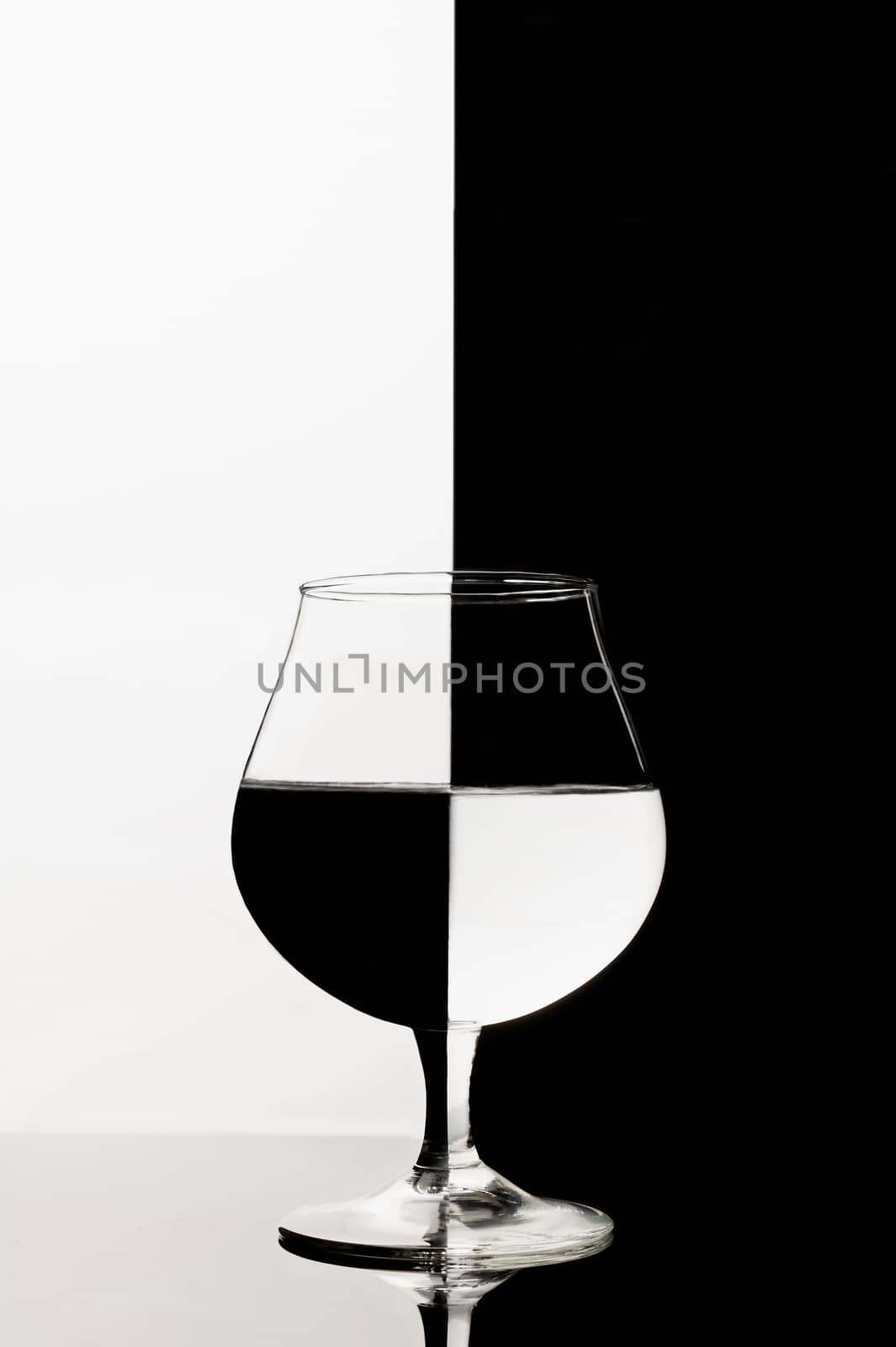A half full cognac glass with interesting refraction effect on black and white background. A half of background is white and other half is black. Stock photo of creative glassware. 