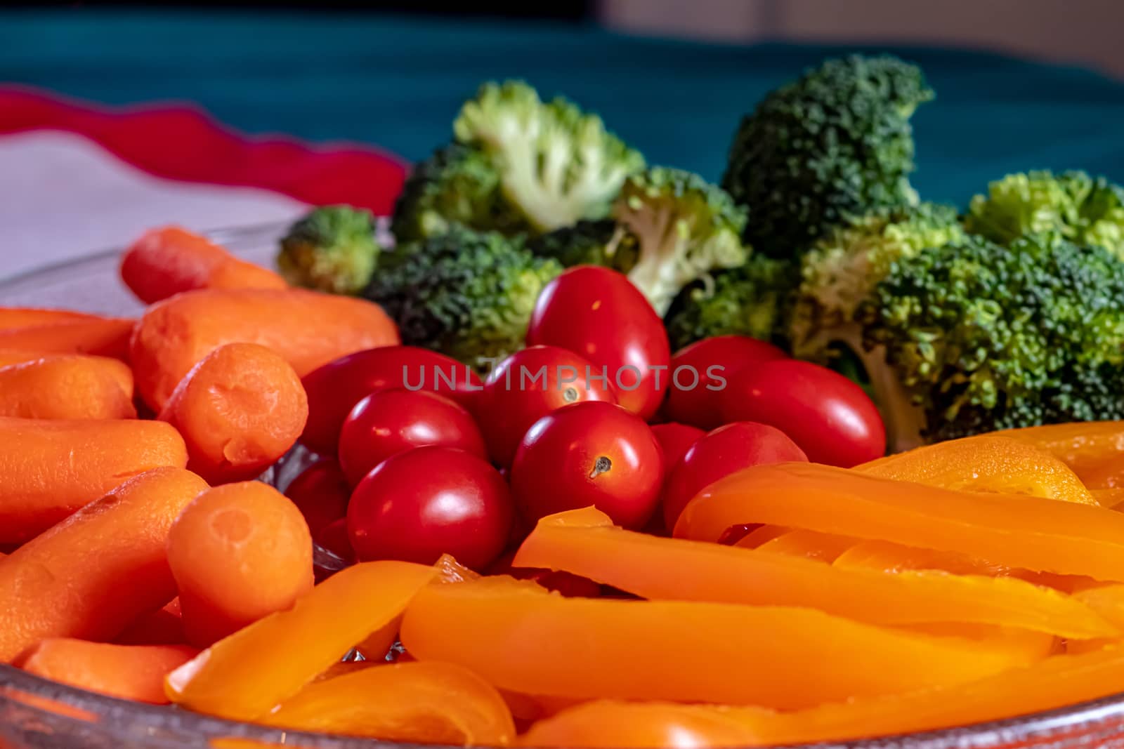 A glass dish of raw veggies consists of broccoli florets, baby carrots, grape tomatoes and sliced yellow bell peppers. The snack tray presents the vegetables on a table with a mat and tablecloth.