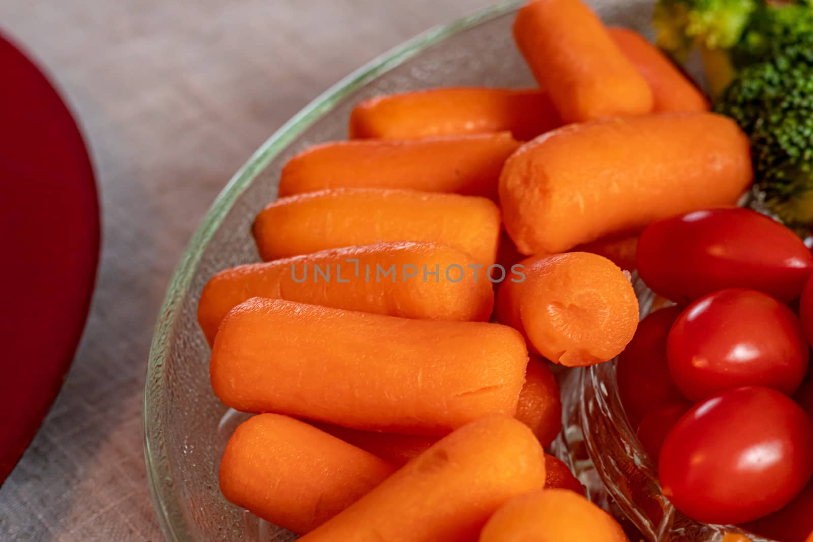 Baby carrots are piled in a glass serving tray with other vegetables. Cut broccoli and grape tomatoes are also seen to their right.