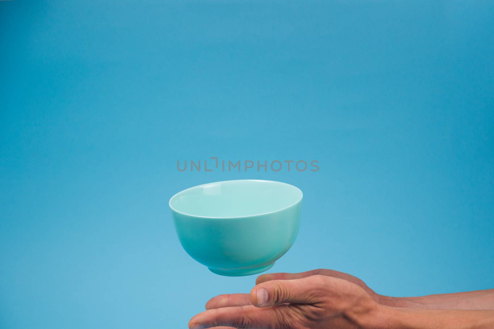 A blue ceramic mattle deep bowl for breakfast flying under male hands on blue background. Ideal photo for levitation of food and fruits or nuts.