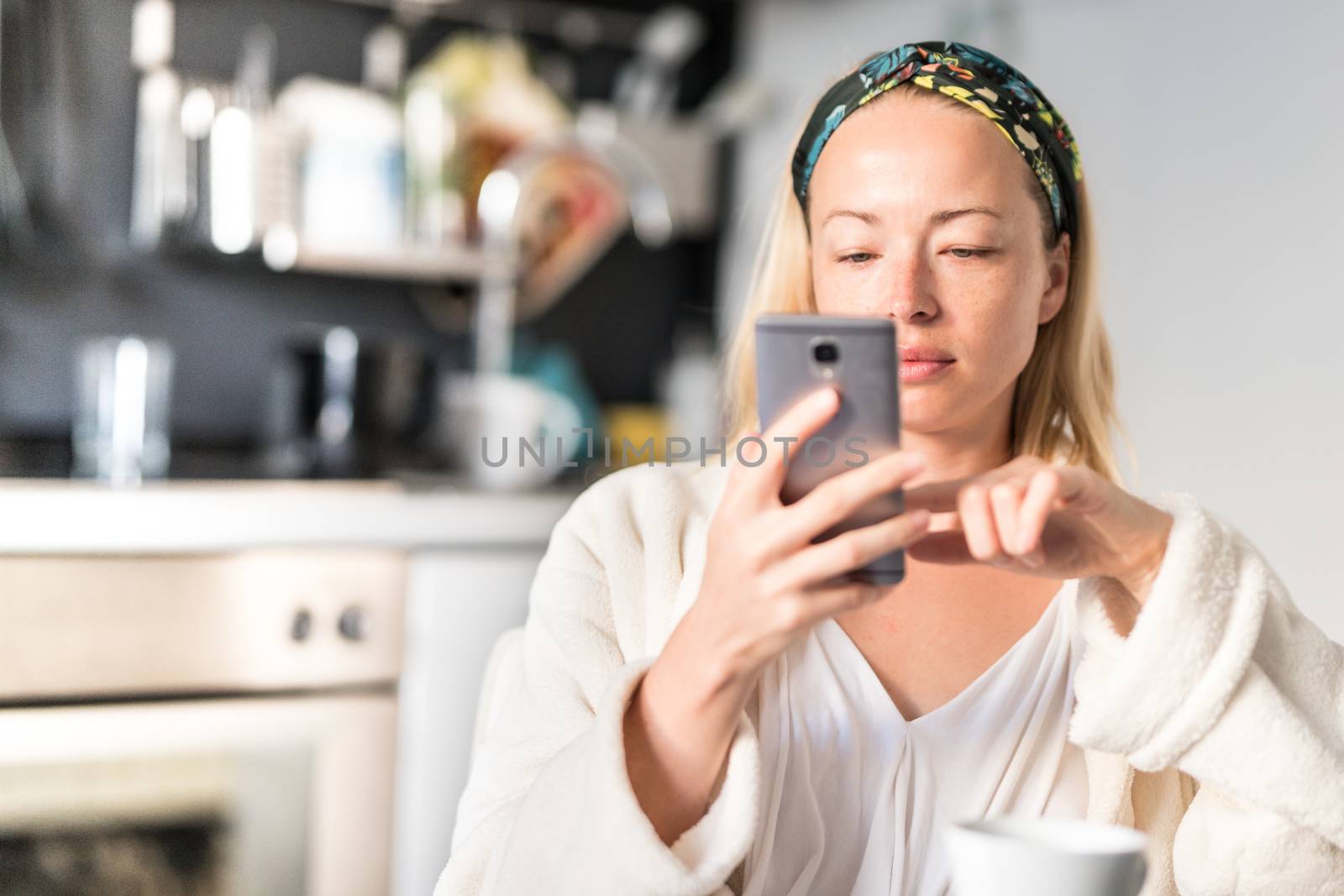 Beautiful caucasian woman at home, feeling comfortable wearing white bathrobe, taking some time to herself, drinking morning coffee and reading news on mobile phone device in the morning.