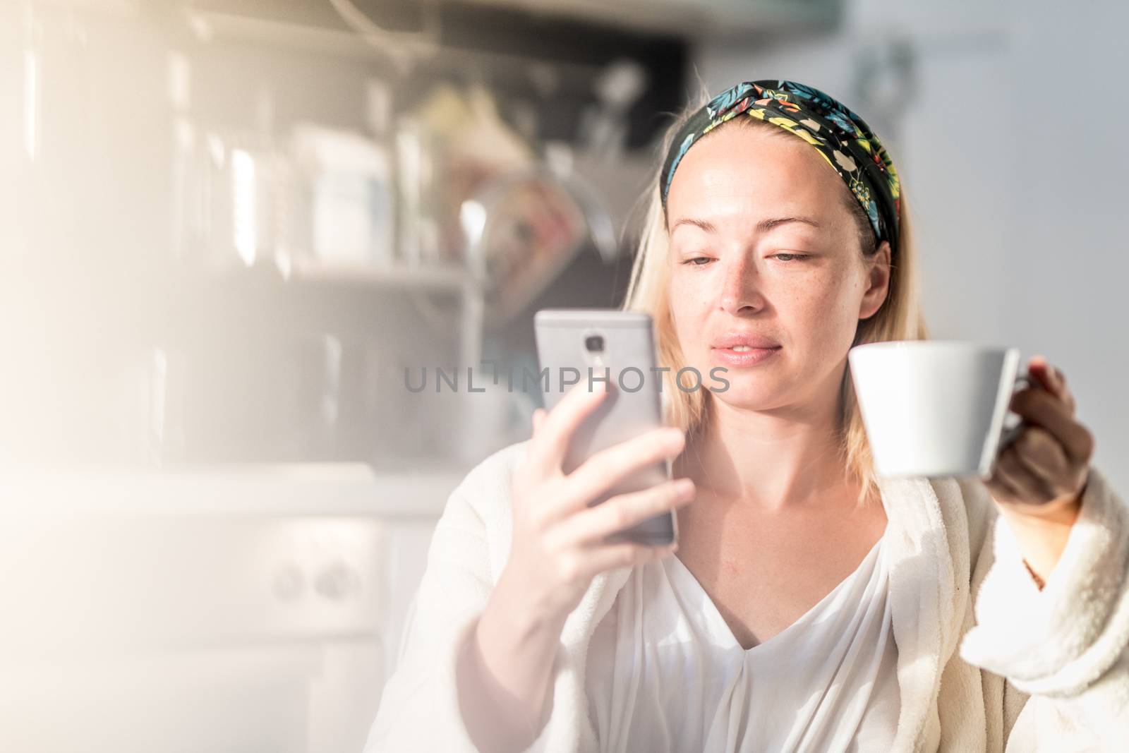 Beautiful caucasian woman at home, feeling comfortable wearing white bathrobe, taking some time to herself, drinking morning coffee and reading news on mobile phone device in the morning.