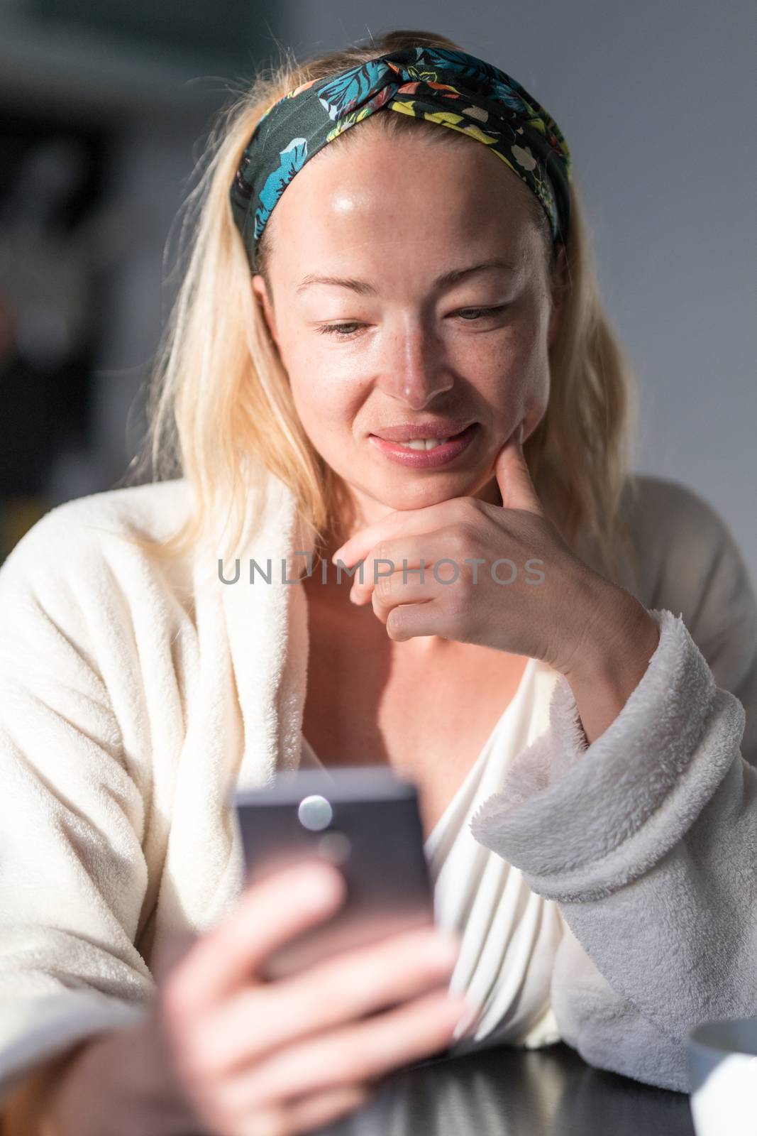 Beautiful caucasian woman at home, feeling comfortable wearing white bathrobe, taking some time to herself, drinking morning coffee and reading news on mobile phone device in the morning.